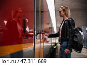 Young blond woman in jeans, shirt and leather jacket wearing bag and sunglass, presses door button of modern speed train to embark on train station platform. Travel and transportation. Стоковое фото, фотограф Matej Kastelic / Фотобанк Лори