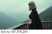 A woman with a sleek gray bob dressed in a black ensemble gazes pensively from a balcony, surrounded by fog-covered mountains. The atmosphere is serene yet introspective. Стоковое AI-изображение, AI-художник Sergey Grishin / Фотобанк Лори