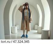 A woman stands confidently in a stylish beige outfit, showcasing blue sneakers, in a modern, minimalist space defined by soft arches. Стоковое AI-изображение, AI-художник Sergey Grishin / Фотобанк Лори