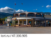 A fragment of the Yalta embankment, one of the most popular resort towns in Crimea. In the foreground is a modern building with a round façade and stained-glass windows, where cafes and restaurants are located. On the left is part of the pedestrian zone with passers-by, vacationers and strollers along the shore. In the background, the green mountains of the Crimean ridge rise, and the sky is decorated with living clouds and a flying bird. The Russian flag on the mast emphasizes the formality and maritime character of the place.The decor conveys an atmosphere of relaxation, resort activity and harmony with the natural landscape. (2021 год). Редакционное фото, фотограф Анна Кривицкая / Фотобанк Лори
