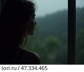 A reflective woman stands by a window, watching the rain fall gently outside. The lush greenery and soft light create a serene atmosphere, evoking a sense of calm and introspection Generative AI. Стоковое AI-изображение, AI-художник Юлия Петровская / Фотобанк Лори