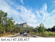 A picturesque section of the highway on the southern coast of Crimea. Ahead, a majestic view of the Kush-Kaya rock massif rises above the green forest. Cars and construction equipment move along the road. On both sides of the highway there is dense vegetation, trees, and tall grass. The sky is clear, with light cirrus clouds emphasizing the scale of the mountains and the vastness of the southern landscape. The scene conveys a sense of freedom, nature, and travel along Crimean routes. (2021 год). Редакционное фото, фотограф Анна Кривицкая / Фотобанк Лори
