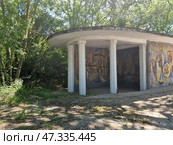 An old Soviet bus stop, lost in the green forest area of ​​Crimea. The structure has a characteristic round roof supported by white columns and is decorated with a mosaic panel. The mosaic depicts abstract and symbolic scenes - human figures, architectural elements, scales, wheels and signs referring to mythology, history and labor are recognizable. The panel is made in bright but faded colors. The state of the stop speaks of neglect: garbage on the ground, a bottle by the column, cracks in the concrete. Around - a thick shadow from the trees, giving the place an atmosphere of solitude and time that seems to have stopped. Стоковое фото, фотограф Анна Кривицкая / Фотобанк Лори