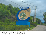 Entrance sign of the resort complex "Ai-Danil", located in Crimea. The sign is made in a modern style: a large decorative structure in the form of a ring, made in beige and blue tones, inside which is the silhouette of a sailboat - a symbol of sea recreation and travel. The inscription reads: "Ai-Danil - a health resort complex". The sign is installed against the background of a green wooded slope, along the road going towards the mountains. In the frame - a cloudy sky and power lines, emphasizing the remoteness and naturalness of the area. Стоковое фото, фотограф Анна Кривицкая / Фотобанк Лори