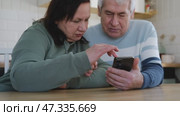 Old couple using a phone together while relaxing in the kitchen at home. Elderly man and woman spending leisure time browsing online with a smartphone. Happy senior married couple typing, learning. Стоковое видео, видеограф Марина Демидюк / Фотобанк Лори