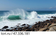 Powerful ocean waves crash onto a rocky coast, creating a splash against the shore under a clear blue sky with a few wispy clouds. The scene reflects a serene atmosphere. Стоковое AI-изображение, AI-художник Sergey Grishin / Фотобанк Лори