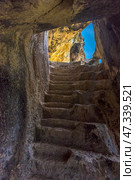 A rock-cut staircase leading up to daylight. The stone steps are uneven, rough, carved by time and probably by human hands. The light pours in from above, creating a beautiful play of shadows and emphasizing the texture of the ancient stone. Such a scene is typical of the cave monasteries of Crimea, especially Chelter-Marmara, one of the most atmospheric cave complexes, located near Bakhchisaray. It was founded in the Middle Ages and was used as a monastery. (2021 год). Стоковое фото, фотограф Анна Кривицкая / Фотобанк Лори