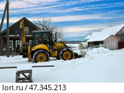 Yellow backhoe uses blade to remove snow in winter rural scene, employing hydraulics and tire traction near rustic wooden buildings under partly cloudy skies. Стоковое фото, фотограф Владимир Григорьев / Фотобанк Лори
