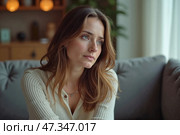 Задумчивая молодая женщина с длинными волосами, сидящая на диване в уютной гостиной. Стоковое AI-изображение, AI-художник Елена Болотина / Фотобанк Лори