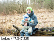 Счастливый 12-летний мальчик держит на руках своего 3-месячного брата. Подросток с младенцем сидит на бревне в весеннем лесу. Стоковое фото, фотограф Яна Проничева / Фотобанк Лори