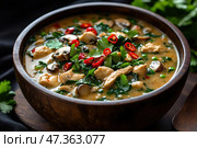 A delicious and aromatic dish, presumably an Asian spicy soup with chicken, mushrooms and herbs. A thick broth filled with pieces of chicken fillet, champignons, green peas, spinach leaves and parsley is served in a wooden bowl. On top are slices of red hot chili pepper, adding piquancy and brightness to the dish. The background is dark, which emphasizes the rich colors of the ingredients. The scene conveys the warmth of home cooking and the rich taste of traditional food. Стоковое AI-изображение, AI-художник Анна Кривицкая / Фотобанк Лори