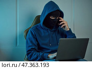 Маскированный хакер работает на ноутбуке в слабо освещенной комнате. Стоковое AI-изображение, AI-художник Елена Болотина / Фотобанк Лори