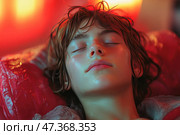 A young man or teenager with his eyes closed, deep in rest or sleep. The skin is slightly damp, perhaps from sweat or water, giving a feeling of coolness or recovery from exertion. The hair is tousled, the face is calm and relaxed. The background is painted in warm red-orange tones, creating an atmosphere of comfort and peace, as if he is in a protective capsule, under the light of a lamp, or inside a futuristic room. The image can be interpreted as a scene of healing, relaxation, meditation, or emotional recovery. Стоковое AI-изображение, AI-художник Анна Кривицкая / Фотобанк Лори