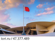 Doha, Qatar - Jan 06 2024, a view of flagpole with the flag of Qatar, fluttering in the wind, with the National Museum of Qatar on background, at evening with cloudy sky, Doha, Qatar. Редакционное фото, фотограф Сергей Богомяко / Фотобанк Лори