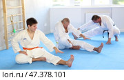 Concentrated adolescent jiu-jitsu fighter doing stretching exercises on mat, warming up body muscles before training during group martial arts class. Стоковое видео, видеограф Яков Филимонов / Фотобанк Лори