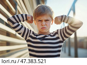 Kid boy shows his muscles. Angry little child boy waves his fist at the camera outdoors at the city street. Unhappy grumpy boy showing his biceps. Provocation. Elementary school problems. Bullying. Стоковое AI-изображение, AI-художник Марина Демидюк / Фотобанк Лори