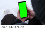 Young man sitting and holding smartphone with green screen outdoors in winter. Vertical mockup of mobile phone. Guy using phone with blank green display background. Smartphone with chromakey. Close-up. Стоковое видео, видеограф Евгений Демидюк / Фотобанк Лори