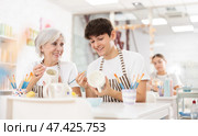 Cheerful guy talking with female colleague while painting mugs in pottery workshop. Стоковое фото, фотограф Яков Филимонов / Фотобанк Лори