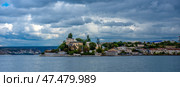 One of the picturesque corners of Sevastopol - a view of the historical buildings along the coast. In the foreground is a calm sea surface reflecting the cloudy sky. In the center of the frame stands out a building with a tower and columns, surrounded by trees and greenery, which is probably one of the historical or military sites of the city. In the distance, old buildings, residential buildings and port cranes are visible, hinting at the industrial and naval significance of this part of Sevastopol. Dark clouds above the horizon create a dramatic atmosphere. Стоковое фото, фотограф Анна Кривицкая / Фотобанк Лори