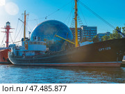 Museum ship SRT-129, moored at the embankment next to the Planetarium of the Museum of the World Ocean in Kaliningrad. It is an old fishing trawler with a black hull and yellow masts, carefully restored for museum purposes. In the background is the spectacular globe-shaped planetarium ball and modern buildings. To the right are reflections of sunlight on the water. To the left of the ship, a bright red lighthouse ship is partially visible. The scene is full of marine and scientific symbolism, and the day is clear and sunny. (2020 год). Редакционное фото, фотограф Анна Кривицкая / Фотобанк Лори