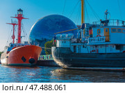 A striking and recognizable scene in Kaliningrad is the museum complex on the embankment. In the foreground are two ships: a red lighthouse ship and a black and white fishing vessel "CPT-129". In the center of the frame, against the backdrop of the ships, rises a spherical building - this is the Museum of the World Ocean with its famous dome-planetarium, lined with mirrored glass. Around - modern urban architecture, water and a clear blue sky. (2020 год). Редакционное фото, фотограф Анна Кривицкая / Фотобанк Лори
