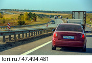 A busy section of highway in an autumn landscape. In the foreground is a Chevrolet Lacetti with a trailer moving along the road. A dividing metal fence is visible, and in the oncoming lane is a stream of trucks. In the distance, the highway smoothly disappears into the horizon, skirting the hills. To the left are soft hills with golden vegetation and sparse trees. Bright daylight and a light haze create the atmosphere of a quiet country trip. (2020 год). Редакционное фото, фотограф Анна Кривицкая / Фотобанк Лори