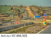 Active construction of a new lane on a country highway. On the left is the existing roadway with heavy traffic of cars and trucks. On the right is a section of the road under construction: equipment, embankment, construction materials and workers are visible. The road passes through hilly countryside with fields, greenery and sparse trees. There are temporary road signs, fences and barriers. Bright sunlight highlights all elements of the scene, creating a sense of scale and movement. (2020 год). Редакционное фото, фотограф Анна Кривицкая / Фотобанк Лори