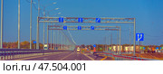 A multi-lane section of highway with perfectly smooth asphalt and metal guardrails. Above the roadway, structures with blue road signs are installed, indicating the directions of movement along the lanes. Light poles with identical curved consoles are built in perspective along the road, creating a rhythmic visual effect. On the right side of the frame is an exit from the highway leading to a parking lot and roadside infrastructure. Cars and trucks are visible in the distance. On the left is an autumn forest and a sign with the mileage. (2020 год). Редакционное фото, фотограф Анна Кривицкая / Фотобанк Лори