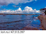A picturesque view of Sevastopol Bay with a concrete embankment and the steps of an amphitheater facing the sea. Along the shore there is a white building with a terrace, and in the distance you can see ships and cranes of shipyards. The weather is sunny but cloudy, creating a striking contrast between the blue sky, sea and architecture. On the horizon you can see the cityscape of Sevastopol with its historical and industrial sites. Стоковое фото, фотограф Анна Кривицкая / Фотобанк Лори