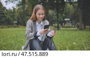 A young girl makes a purchase with her phone and bank card in the park, sitting on the grass. Стоковое видео, видеограф Mikhail Davidovich / Фотобанк Лори