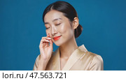 A cheerful Japanese woman showcases her flawless skin, pointing at her face against a blue background, symbolizing the essence of Asian skincare practices. Стоковое AI-видео, аниматор Ирина Старикова / Фотобанк Лори