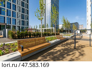 Benches line the pathway in a contemporary urban park, surrounded by tall buildings. Lush greenery and blue skies create a peaceful atmosphere for relaxation and socializing. Стоковое фото, фотограф Алексей Климкин / Фотобанк Лори