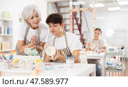 Elderly female teacher assisting guy painting ceramic mug in pottery class. Стоковое фото, фотограф Яков Филимонов / Фотобанк Лори