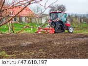 A tractor equipped with a red rotary cultivator actively prepares the soil in a rural field. The equipment loosens the earth, preparing it for planting and cultivation. Стоковое фото, фотограф Коханова Наталья / Фотобанк Лори