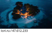 Romantic beachfront cottage illuminated at night surrounded by calm waters and palm trees in a tropical paradise. Стоковое AI-изображение, AI-художник Михаил Коханчиков / Фотобанк Лори