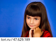 A young girl with striking long hair and bangs poses thoughtfully against a bright blue background. She embodies innocence and charm, capturing the essence of childhood wonder in her expression. Стоковое фото, фотограф Aleksandr Golubev / Фотобанк Лори