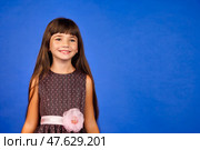 A cheerful girl smiles brightly, wearing a lovely dress adorned with a flower. Her long hair gently frames her face as she radiates joy in a vibrant studio setting filled with creativity. Стоковое фото, фотограф Aleksandr Golubev / Фотобанк Лори