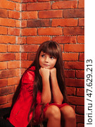 In a charming brick room, a young girl with long hair sits thoughtfully, her red outfit contrasting beautifully with the rustic walls. She rests her chin on her hands, lost in dreams. Стоковое фото, фотограф Aleksandr Golubev / Фотобанк Лори