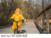 A cute boy in bright yellow clothes rides a bike. A happy child plays outside in early spring. Happy childhood. Стоковое фото, фотограф Photojuli / Фотобанк Лори