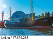 Museum ship SRT-129, moored at the embankment next to the Planetarium of the Museum of the World Ocean in Kaliningrad. It is an old fishing trawler with a black hull and yellow masts, carefully restored for museum purposes. In the background is the spectacular globe-shaped planetarium ball and modern buildings. To the right are reflections of sunlight on the water. To the left of the ship, a bright red lighthouse ship is partially visible. The scene is full of marine and scientific symbolism, and the day is clear and sunny. (2020 год). Редакционное фото, фотограф Анна Кривицкая / Фотобанк Лори