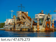 An old industrial transport vessel moored off the coast of Kaliningrad. This is a dredger called "Alexander Uvarov", according to the inscription on the side. The vessel has a massive structure with signs of rust and wear, which gives it an industrial character. It is equipped with cargo and working mechanisms: a conveyor system, manipulators, ladders. In the background is a blue industrial hangar and a mast with the Russian flag. The bright sunny day emphasizes the texture of the metal, water and rust. (2020 год). Редакционное фото, фотограф Анна Кривицкая / Фотобанк Лори