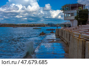 A picturesque view of Sevastopol Bay with a concrete embankment and the steps of an amphitheater facing the sea. Along the shore there is a white building with a terrace, and in the distance you can see ships and cranes of shipyards. The weather is sunny but cloudy, creating a striking contrast between the blue sky, sea and architecture. On the horizon you can see the cityscape of Sevastopol with its historical and industrial sites. Стоковое фото, фотограф Анна Кривицкая / Фотобанк Лори