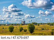 A rural or suburban landscape on a clear day. In the center of the frame rises an industrial building, perhaps a grain silo or factory, surrounded by low buildings. A spacious field with sparse trees stretches in the foreground. The main emphasis is on the sky, which takes up most of the frame and is dotted with beautiful cumulus clouds, creating depth and an aerial perspective. The lighting is soft, with daytime contrast, emphasizing the terrain and the volume of the clouds. Стоковое фото, фотограф Анна Кривицкая / Фотобанк Лори