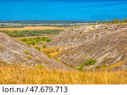 A picturesque view of chalk hills covered with dry steppe vegetation. In the foreground is tall dry grass, typical of the southern Russian steppe. Whitish stripes of chalk and sparse bushes are visible on the slopes of the hills. Below is a green valley with trees, behind it are yellow fields and a village. On the horizon, a forest and a light haze are visible. The bright blue sky without clouds emphasizes the contrast between the hills and the plain. This is probably one of the natural landscapes of the Voronezh region, for example, Divnogorye. Стоковое фото, фотограф Анна Кривицкая / Фотобанк Лори