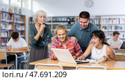 Adult librarian holding his finger on lips making silent gesture in library to laughing teenage pupils watching online video. Стоковое фото, фотограф Яков Филимонов / Фотобанк Лори