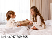 Two sisters wearing white dresses, sitting on a bed with white bedding, pulling big wrapped gift box, fighting, cannot share a present on the holiday morning at home. Стоковое фото, фотограф Ирина Михайличенко / Фотобанк Лори