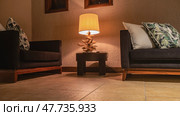 A place to relax in the room. Two sofas with soft patterned cushions against a beige wall in the corner. The desk lamp is lit. Translucent lampshade, designer wooden stand. Evening lighting. Стоковое фото, фотограф Вера Ивановна Кубарева / Фотобанк Лори
