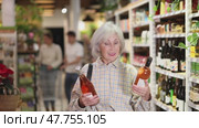 Mature woman chooses Italian wine in the alcohol department of a hypermarket. Adult woman with gray hair choosing semi-sweet wine for dinner. Стоковое видео, видеограф Яков Филимонов / Фотобанк Лори