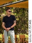 A young man stands in a lush Dubai garden, dressed in casual attire and sunglasses. He holds a stick, embodying a relaxed yet stylish demeanor amidst greenery and outdoor seating. Стоковое фото, фотограф Aleksandr Golubev / Фотобанк Лори