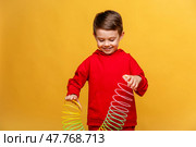 Happy smiling little boy 3-4 years old, wearing fashion red sport suit, plays with a plastic colored neon rainbow spiral children toy, holding bouncy stretchy toy on yellow background, copy space. Стоковое фото, фотограф Ирина Михайличенко / Фотобанк Лори